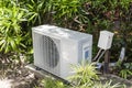 A split type inverter air conditioning condenser mounted outside a house partially concealed with shrubs. HVAC for tropical