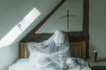 A spooky, ghostly figure sitting on a bed, covered in a sheet with a cross above the bed