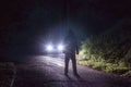 A spooky hooded figure, silhouetted against a cars headlights. On a rainy country road at night