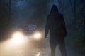 A spooky hooded figure silhouetted against a cars headlights. On a scary forest road at night