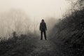 A spooky hooded figure standing on a country path on a eerie foggy winters day