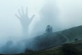 A spooky transparent hooded figure holding up it`s hand. Appearing over a moody hillside. On a foggy winters day in the