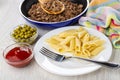 Spoon in frying pan with fried mince, napkin, bowls with green peas, ketchup, plate with pasta, fork on table