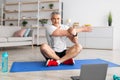 Sport in mature age. Happy middle-aged man doing stretching exercises in front of laptop at home, sitting on mat Royalty Free Stock Photo
