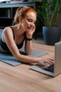 Sporty smiling redhead young woman lying on floor and using laptop computer at apartment. Royalty Free Stock Photo