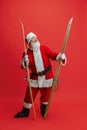 Full-length portrait of senior man wearing Santa Claus costume, holding skis isolated over red background. Winter Royalty Free Stock Photo