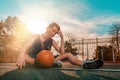 Sports and basketball. A young teenager in a blue tracksuit poses with a ball while sitting on a sports field. Blue sky and