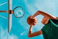 Sports and basketball. A young teenager in a green tracksuit throws a ball into the basket. Bottom view. Blue sky in the