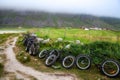 Several sports bikes with thick wheels are parked in a field by the empty country road under the hills. Low clouds. Norway