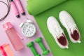 Sports concept. Top view photo of white sneakers over green exercise mat dumbbells alarm clock skipping rope pink bottle and Royalty Free Stock Photo