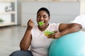 Sports and healthy eating for slimming. Plump black woman with fitness ball enjoying tasty vegetable salad at home