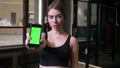 Sportsgirl showing smartphone with chromakey green screen into camera being serious and concentrated in gym.