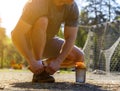 Sportsman laces his sneakers sitting and looking forward. Blurred background with forest, kids playground and football goal