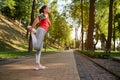 Side portrait of a middle-aged sporty woman stretching her leg muscles during a morning workout in a city park on a sunny warm Royalty Free Stock Photo