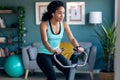 Sporty african young woman exercising on smart stationary bike and listening to music at home