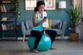 Sporty african young woman using her mobile phone after session of exercises while sitting on fitness ball at home Royalty Free Stock Photo