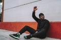 Sporty Black man in black sweaters and jogging pants sitting on concrete pavement outside a building