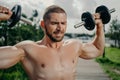 Sporty determined muscular European man exercsises with dumbbells, makes weightlifting outdoor, has naked torso, trains muscles,
