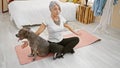 Sporty grey-haired middle-aged woman with pet dog, training yoga in serene bedroom ambiance, sitting on floor near bed for healthy
