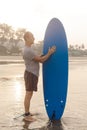 Sporty man with leash attached to ankle keeping hand on surfboard and looking at equipment on sandy seashore at sunset.