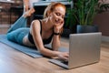 Sporty smiling redhead young woman lying on floor and using laptop computer at apartment. Royalty Free Stock Photo