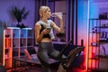 Sporty woman using mobile phone, drinking water while riding on exercise bike at home Royalty Free Stock Photo