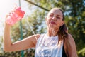 Sporty young blonde woman with sporty glass with cool illuminating water pours water on herself on sports field and laughs