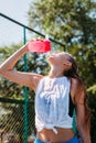 Sporty young blonde woman with sporty bottle with cool water pours water on herself on sports field