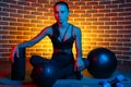 Sporty young brunette woman posing with medicine balls and foam fitness roller on mat in gym in neon lights.