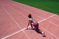 A sporty young dark-skinned girl in a gray T-shirt, pink pants and sneakers stands on one knee on the starting line of the stadium Royalty Free Stock Photo