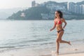 Sporty young woman in bikini running along the beach with bright sunlight and mountainous resort city in background Royalty Free Stock Photo