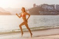 Sporty young woman in bikini running along the beach with bright sunlight and mountainous resort city in background Royalty Free Stock Photo