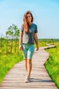 A sporty young woman walks along a picturesque wooden walking path through a swamp with tall grass in summer. Royalty Free Stock Photo