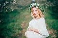 Spring coming! Beautiful young cheerful pregnant woman in wreath flowers on head touching belly while walking in spring tree garde Royalty Free Stock Photo
