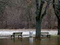 Spring flood. The river has overflowed in the city park, and benches and a trash can are in the water. Very gloomy weather