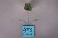 Spring forward sign handwritten on blue posted sticky note with small house desk plant and paper clips shape of flower