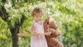 Spring and happy summer time. Joyful smiling little girl playing with teddy bear swinging on the swing. Child with blue eyes and Royalty Free Stock Photo