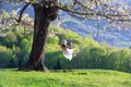 Spring landscape of cherries covered with flowers and green leaves. A girl in a hat, white dres swings under a tree on a swing