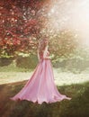 Spring woman walks in blooming sakura tree garden. Medieval long-haired girl, mysterious blonde lady in long pink satin Royalty Free Stock Photo