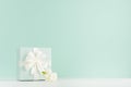 Springtime festive background with white freesia flowers, standing closed square gift box with ribbon on green mint menthe.