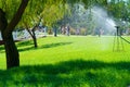 Sprinklers water the green lawn in the city park on a summer day, people walking and children playing, bright sunlight and shadows