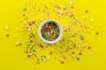 Sprinkles on yellow background with single little white bowl with multi-colored sprinkles and star shapes in centre - top view