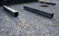 Square with park benches made of natural oak wooden prisms. trunk cut into a block shape. granite paving and stone walls. trees in