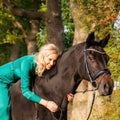 Square portrait of a blonde girl in a vintage green dress with a big skirt posing with a brown horse. Selective focus