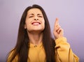 Squint smiling young desiring woman in yellow hoodie crossing fingers with closed eyes on purple bright background. Closeup