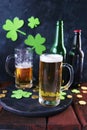 St. Patrick`s Day, fresh beer in glass mugs and a bottle, gold coins, on a red wooden table, a green shamrock on a dark background Royalty Free Stock Photo