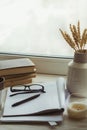 A stack of books, cup of coffee, bouquet of kernels, glasses, notebook and pen over a window.