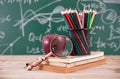 A stack of books in front of a blackboard full of mathematical formulas, a red apple, a magnifying glass and a pen holder filled w