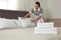 Stack of clean towels with flower on bed and blurred chambermaid in hotel room. Space for text