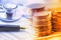 Stack of coins with a fountain pen and an chest-piece of a stethoscope on a table.